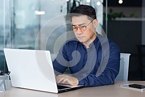 Serious thinking asian businessman working inside modern office, mature man in shirt and glasses using laptop at work