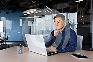 Serious thinking asian businessman working inside modern office, mature man in shirt and glasses using laptop at work