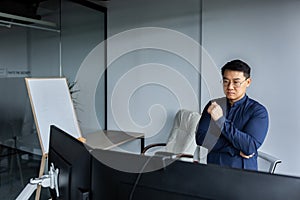 Serious and thinking asian engineer looking at two monitors, close-up photo of office worker, man working inside modern