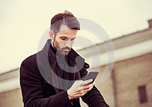 Serious thinking business man in fashion clothing texting sms looking on mobile phone in the hand outdoors autumn background