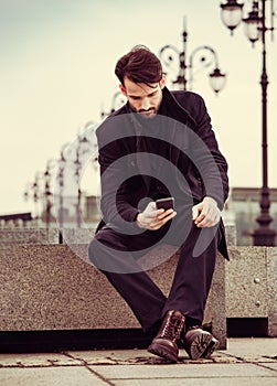 Serious thinking business man in fashion clothing texting sms looking on mobile phone in the hand outdoors autumn background