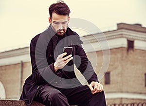 Serious thinking business man in fashion clothing texting sms looking on mobile phone in the hand outdoors autumn background