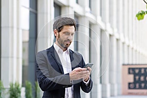 Serious thinking businessman boss holding phone, man reading online message and typing, investor outside office building