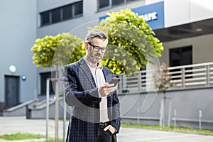 Serious thinking and focused mature businessman using app on phone, senior boss holding smartphone walking around town