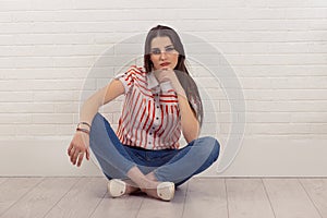 Serious thinking young woman sitting cross-legged on floor, touching chin, looking at camera, isolated white brick wall background