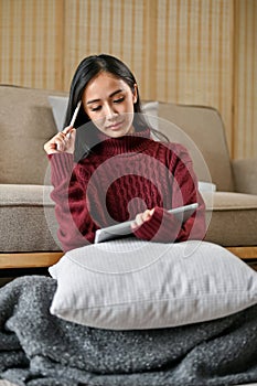 A serious and thoughtful Asian woman in a cozy sweater works on her tasks on her tablet