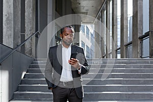 Serious and thoughtful businessman reading online news on phone african american boss in suit outside office building