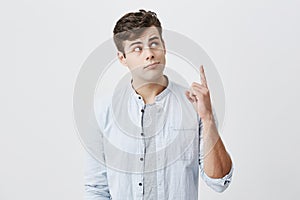 Serious thoughtful european man in light blue shirt over gray t-shirt looking upwards, pointing index fingers up with