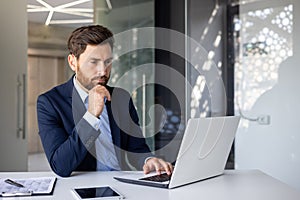 A serious and thoughtful young man businessman is sitting in the office at the desk and is concentrating on his laptop