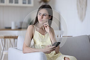 Serious thoughtful young woman holding smartphone oh home couch