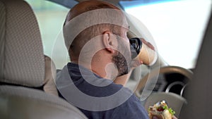 Serious tired man eating fast food and drinking coffee while driving a car. The taxi driver took a break on lunch in