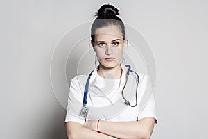 Serious, tired young doctor with a stethoscope in a white coat, standing with arms crossed and looking at the camera
