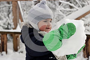 Serious toddler boy in warm coat and green mitts holding a huge snowball and having fun in forest