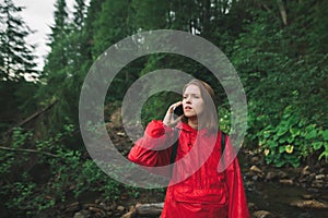 Serious tourist girl speaking on the phone looking far ahead in the mountain fir forest. Female hiker in red raincoat is making a