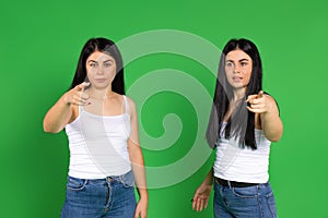 Serious twin sisters in the same clothes point in front with a forefinger on a green background.