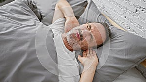 Serious unhurt middle age man, with grey hair and glasses, distressed by cervical pain, lays awake, touching neck in bed - a