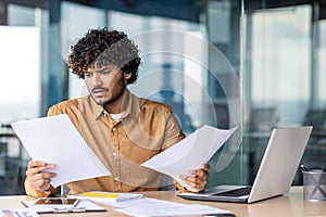 Serious and upset businessman checking and analyzing documents financial reports, thinking hispanic man reviewing