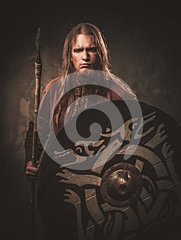 Serious viking with a spear in a traditional warrior clothes, posing on a dark background.