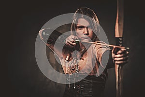 Serious viking woman with bow and arrow in a traditional warrior clothes, posing on a dark background.