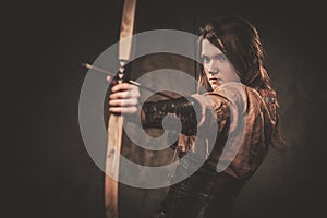 Serious viking woman with bow and arrow in a traditional warrior clothes, posing on a dark background.