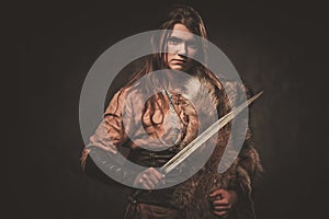 Serious viking woman with sword in a traditional warrior clothes, posing on a dark background.