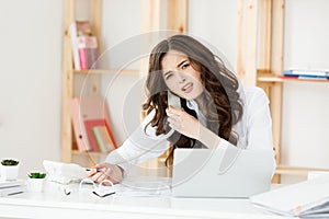 Serious well-dressed saleswoman talking on phone in office behind her desk and laptop computer. Copy space