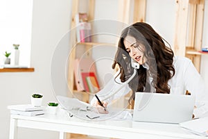 Serious well-dressed saleswoman talking on phone in office behind her desk and laptop computer. Copy space