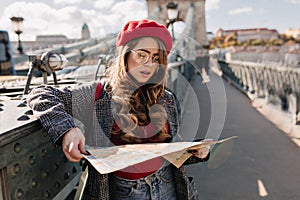 Serious white female traveler looking at map standing on city background. Outdoor photo of woman lost in foreign town