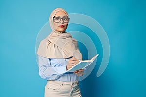 Serious woman in hijab and glasses with pen and book posing on blue background with copy space
