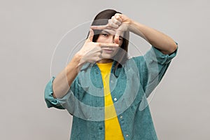 Serious woman looking attentively through photo frame shape with fingers, making photography gesture
