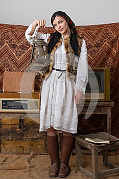 Serious woman in national ukrainian clothes and leather old shoes stands among retro, vintage room, holds kerosene lamp