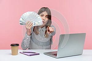 Serious woman office worker holding hundred dollar bills and pointing finger at camera sitting at workplace, making you to earn