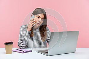 Serious woman office worker talking phone typing on laptop, receiving calls and making online order, coffee cup and notepad lying