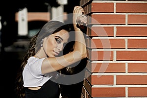 A serious woman in profile leaning against a brick wall wearing makeup and jewelry and dressed in casual clothes