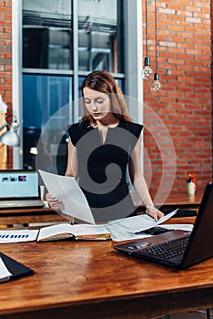 Serious woman reading papers studying resumes standing at work desk in stylish office