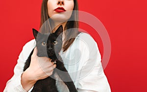 Serious woman with red lips and in a white shirt holds a black cat in her hand. Cat sits in the hands of a girl and looks at the