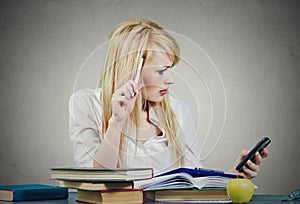 Serious woman sitting at desk with a pile of books , reading a message on her cellphone