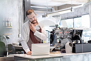 Serious worried barista man taking a call on his mobile phone  checking information received on his laptop with a serious