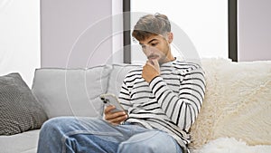 Serious young arab man doubting his next idea, deeply immersed while texting on his smartphone, sitting solo on the living room