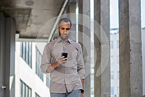 A serious young Arab man in a shirt walks outside an office building and uses a mobile phone intently