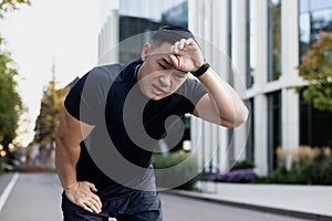 Serious young Asian male athlete and runner wiping sweat from forehead, suffering from headache, bent over from fatigue