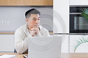 serious young asian man looking away. student writer sit at home office desk with laptop thinking of inspiration search