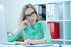 Serious young attractive business woman in glasses sitting at office chair working at desktop computer talking on mobile