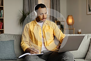 Serious young black man in headphones having online business meeting on laptop, writing down info at home