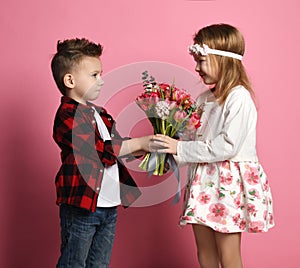 Serious young boy in blue jeans and red shirt hands a spring bouquet to a little girl in pink dress and wreath of flowers