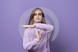 Serious young brunette woman showing time out sign, wears purple hoodie, standing over purple background. Concept of warning,