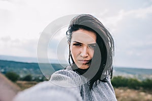 Serious young brunette woman taking self portrait and posing against meadow and overcast sky background. Cute female take selfie