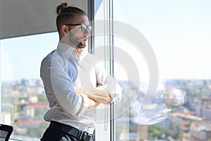 Serious young business man keeping arms crossed and looking away while standing near the window