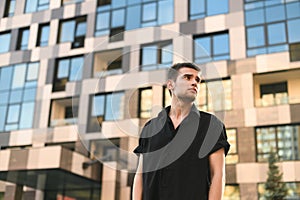 Serious young business man standing on urban background. Guy model in black shirt stands against the background of a modern