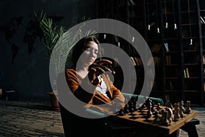 Serious young business woman in elegant eyeglasses making chess move sitting on armchair in dark library room, on